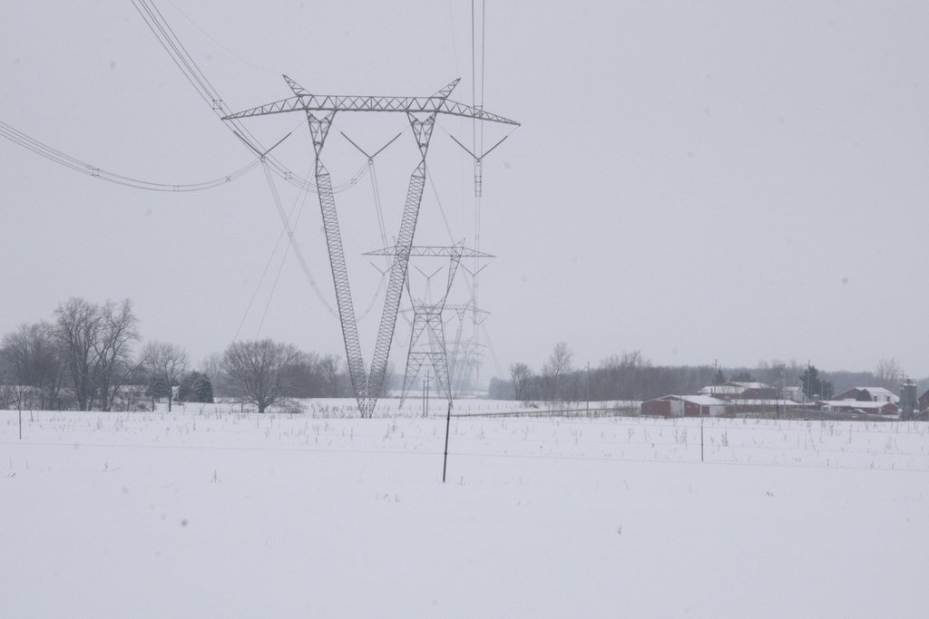 A 765 kV transmission line crossing snow-covered farm fields: a row of very large guyed-V lattice towers receding to the horizon past a farmstead.