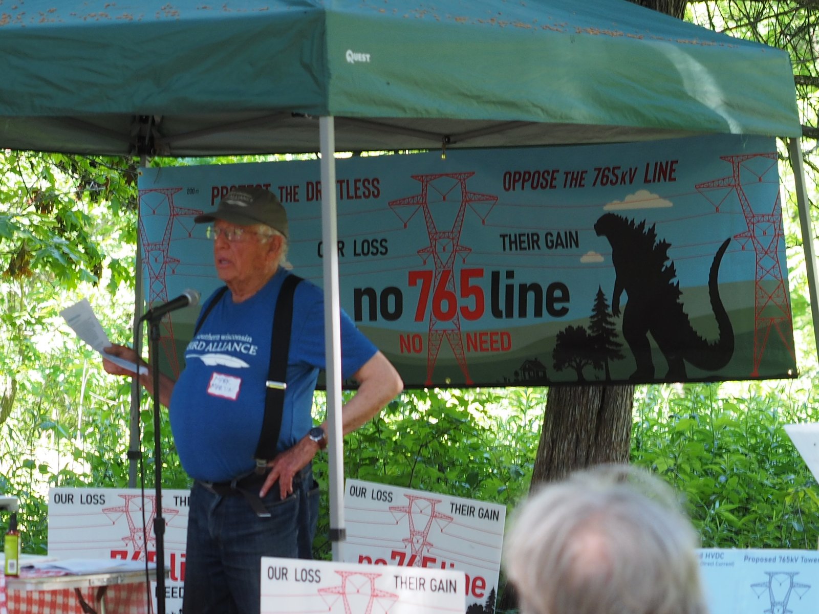 A speaker addresses neighbors at a campaign field event beneath the No 765 Line banner, with yard signs on the table.