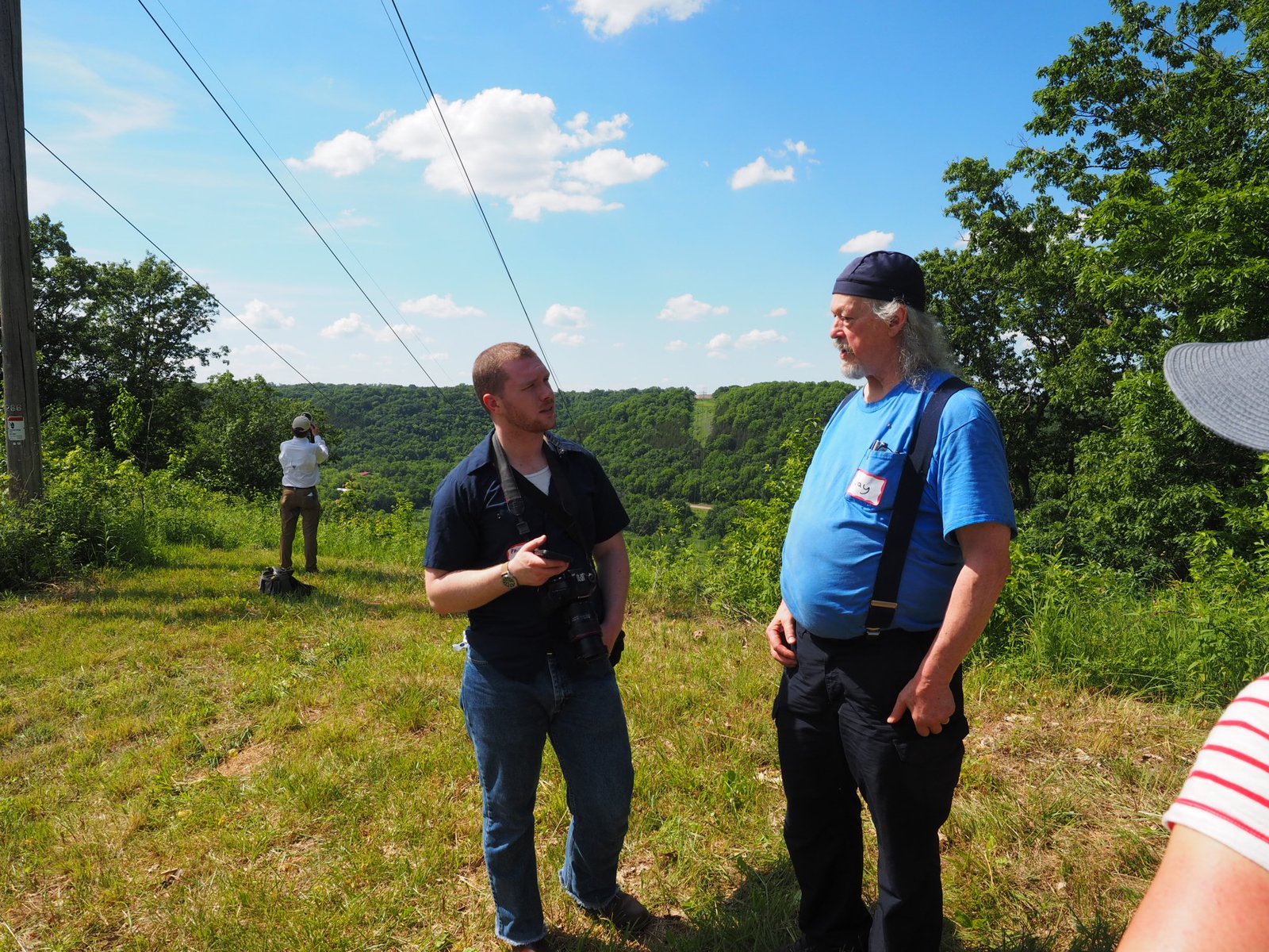 Two neighbors talk on a Driftless hilltop beneath the existing transmission line, the river valley spread out behind them.