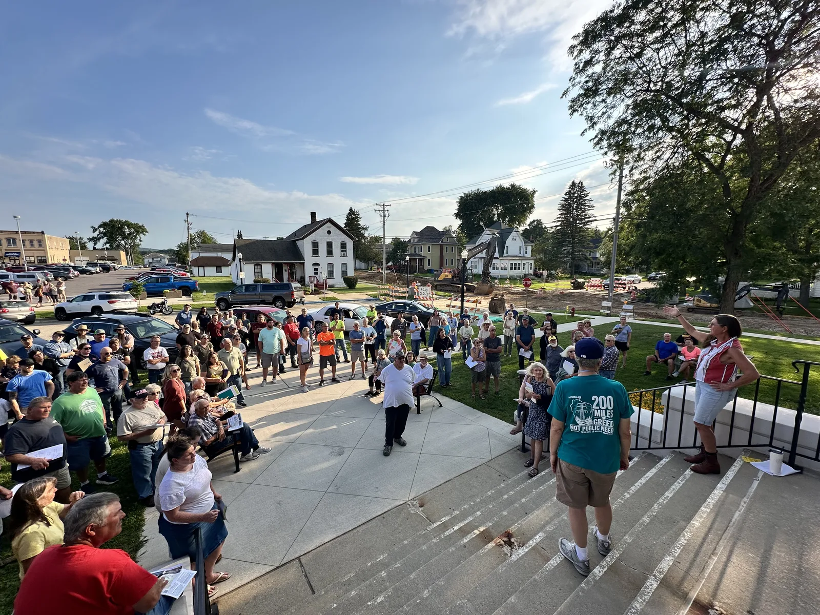A crowd of well over a hundred people gathered on the plaza and lawn outside the Baraboo Public Library on a sunny evening, many holding papers, as a woman addresses them from the top of the entrance steps; one attendee's shirt reads 200 Miles of Greed, Not Public Need.