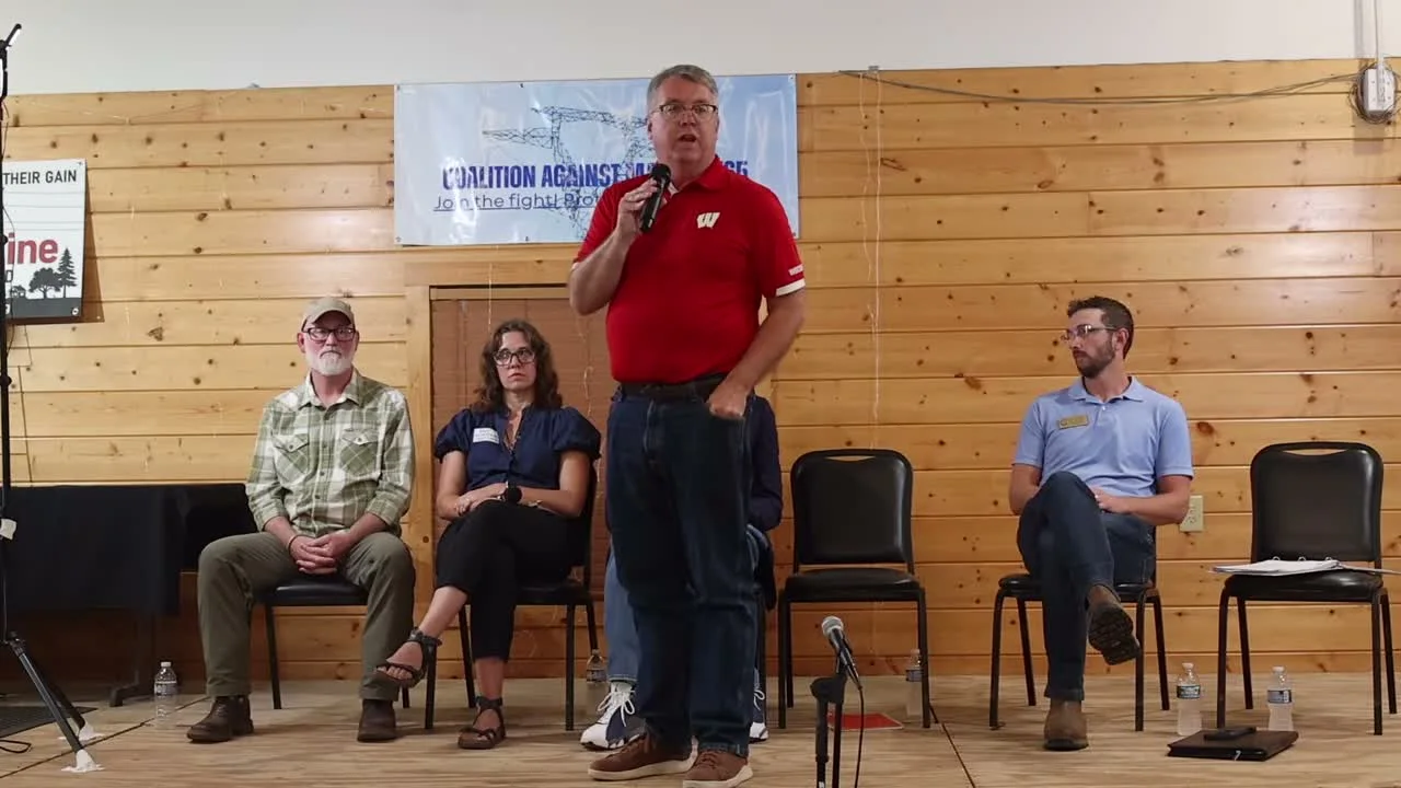 Sen. Brad Pfaff, in a red polo, stands speaking into a microphone; Rep. Derrick Van Orden, Mayor Krista Browne, and Rep. Travis Tranel are seated behind him on the stage.